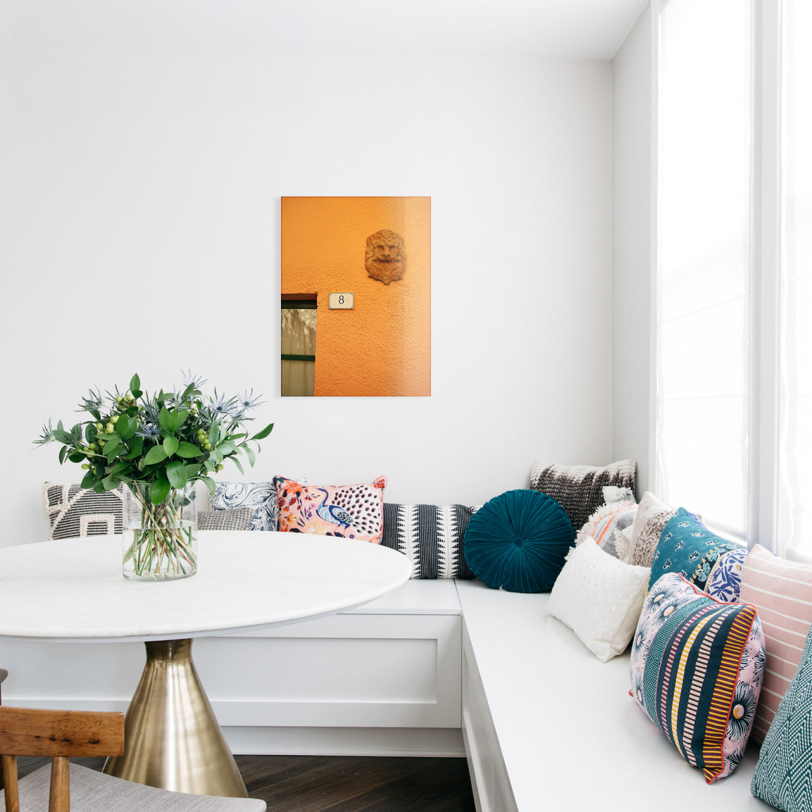 Kitchen nook with a white bench, decorative pillows, a small table with flowers, and a glass print of an orange wall with a carved lion's head and number 8 sign.
